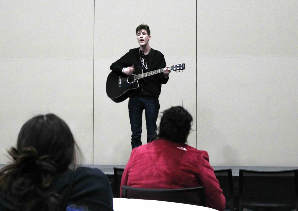 E.R. standing in front of the audience playing a song on the guitar and singing
